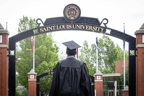 A student wearing a mortarboard stands in front of the Saint Louis University pedestrian gate