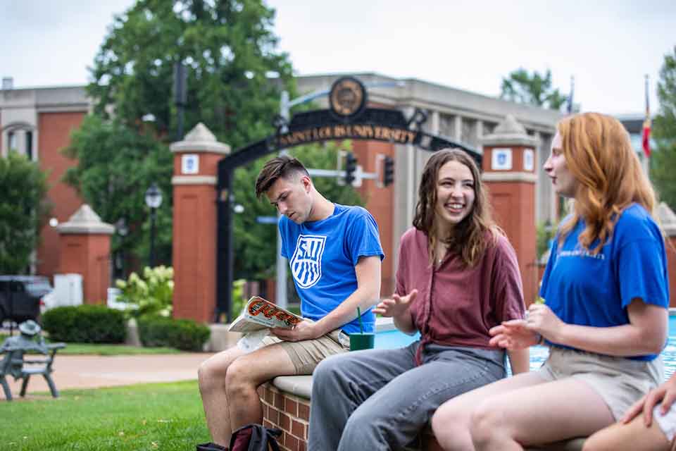 Students sit on the edge of the dolphin pond with Grand Blvd and the Saint Louis University gateway in the background on a sunny day