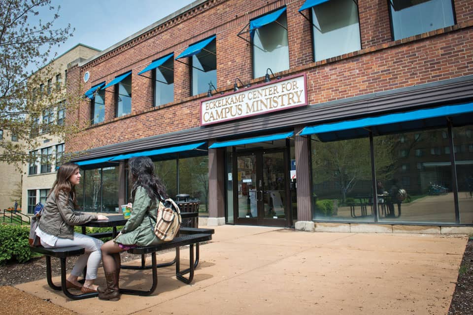 Two people sit at a picnic table outside Eckelkamp Center for Campus Ministry on a sunny day.