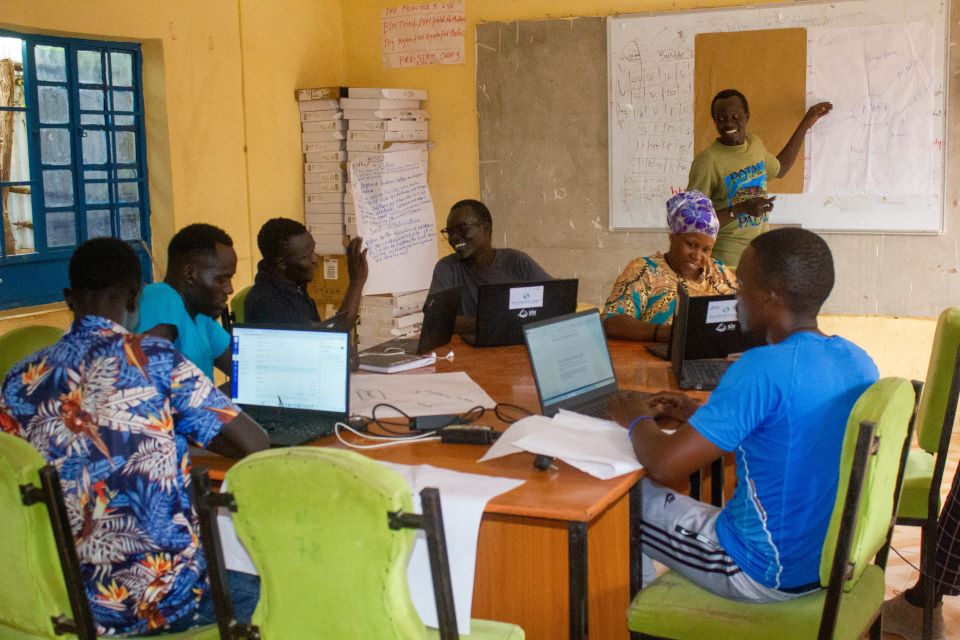 A study group sit around a table. Laptops are on the table in front of them. A man stands in the background pointing at a board.