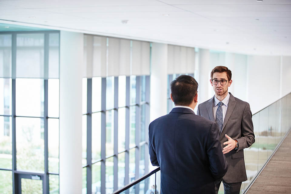 Two male students in suits standing against a rail, having a conversation. 