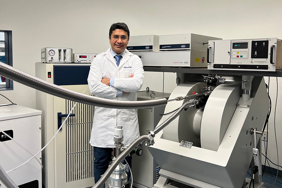 Reza Dastvan, Ph.D., in his lab, wearing a lab coat and standing next to some large pieces of equipment.