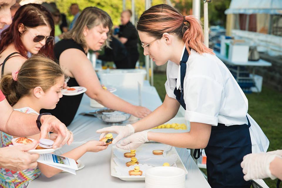 A student wearing an apron reaches over a table to hand a piece of food to a child.